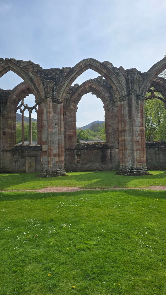 Part of the ruined nave at Melrose Abbey. Two rows of high stone archways with different decorative stone work inset frame a lush green landscape.