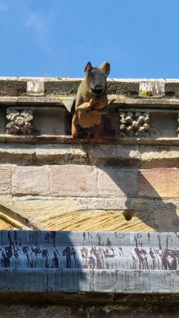 A stone figure of a pig holds bagpipes. The stone gargoyle is set high up on a stone wall.