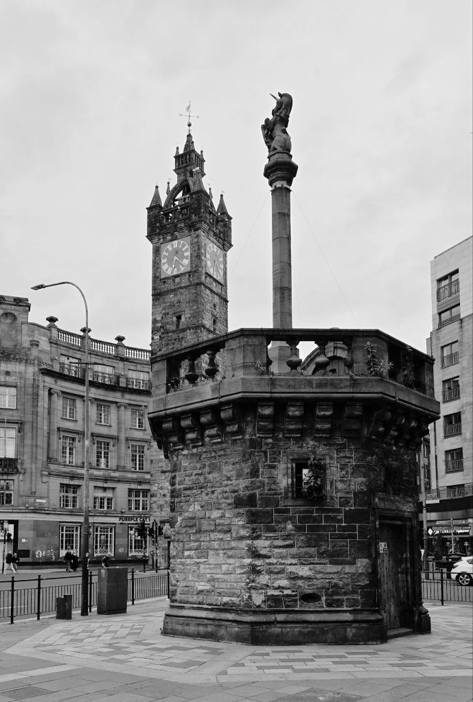 Mercat Cross of Glasgow city centre. Octagonal stone base with long, slender podium featuring a unicorn holding a shield. The clock tower is in the background with a mixture of Playfair-esque and contemporary architecture.