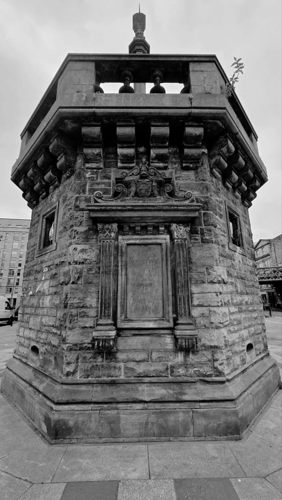 A wide and front-on look at Glasgow's mercat cross octagonal base. Fancy stone frame featuring the text 'Glasgow Flourish' and a plaque commemorating the date the mercat cross was placed are central.