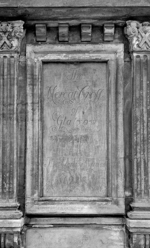 Framed plaque featured on Glasgow's Mercat Cross. The rectangular stone plaque is flanked by carved columns atop which are a St. Andrew's Cross. The plaque reads 'The Mercat Cross of Glasgow, Built in the Year of Grace 1929'